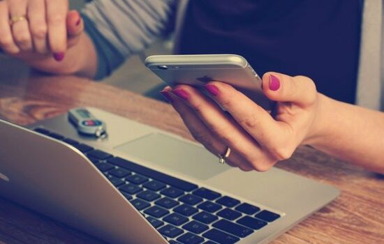 Eine Frau am Laptop mit einem Smartphone in der Hand. der Kopf ist abgeschnitten.