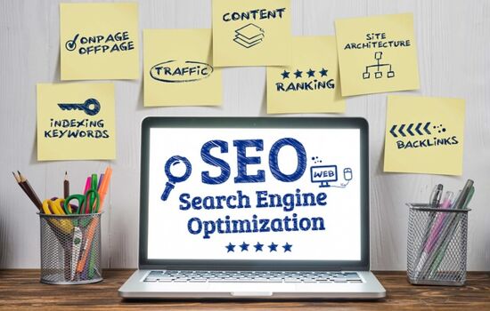 Laptop auf einem Tisch. Auf dem Bildschirm steht SEO Search Engine Optimization. An der Wand über dem Laptop kleben Post-Its mit INDEXING KEYWORDS, ONPAGE/OFFPAGE,TRAFFIC, CONTENT, RANKING, SITE ARCHITECTURE und BACKLINKS